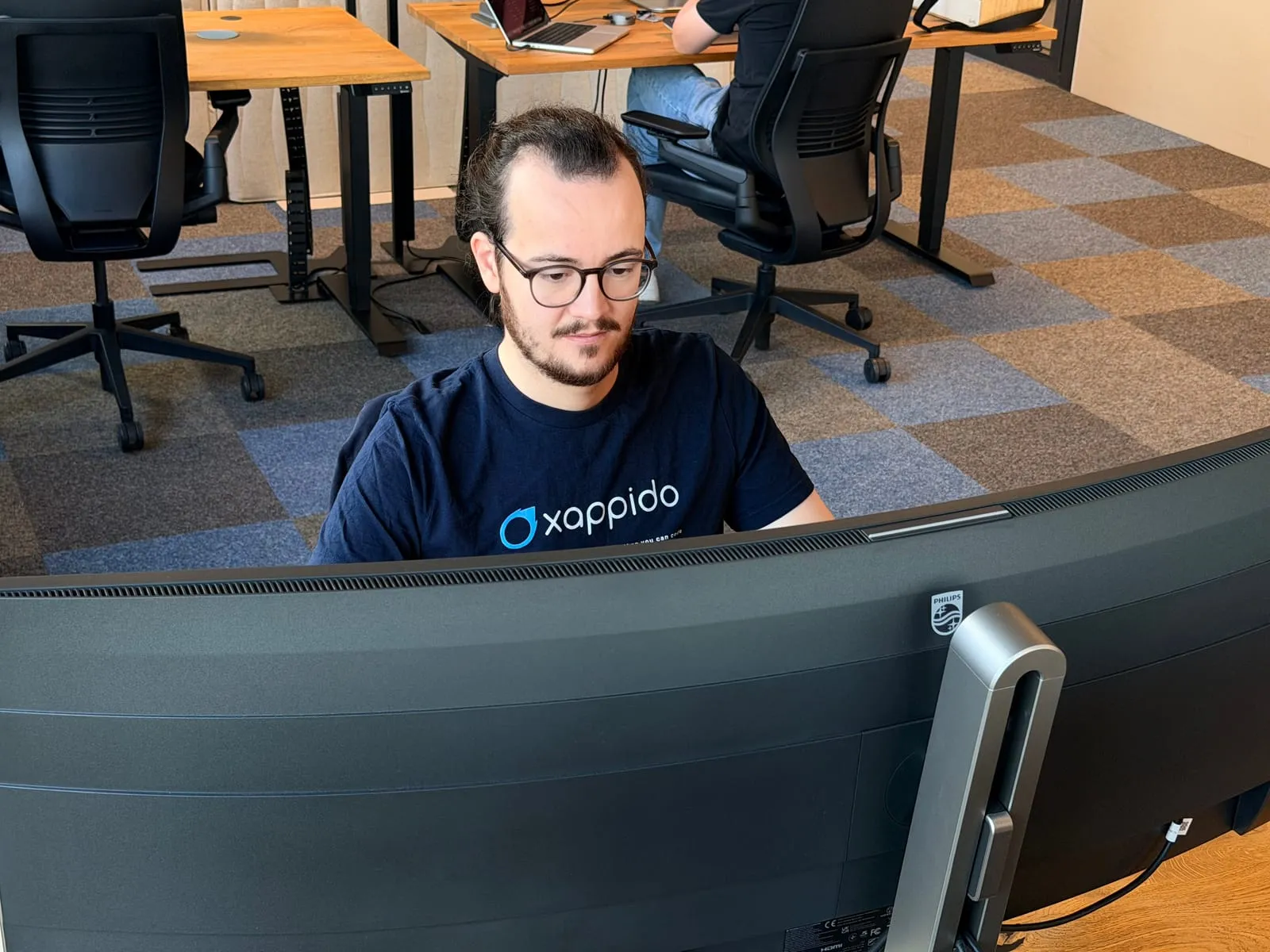 Pablo arbeitet hinter einem breiten, gebogenen Bildschirm im Grossraumbüro