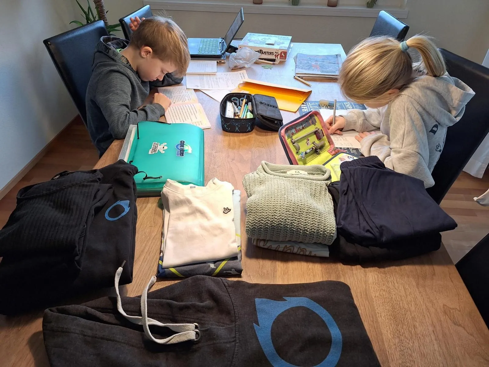 Zwei Kinder machen am Esstisch Hausaufgaben, davor liegen Schultaschen und Jacken