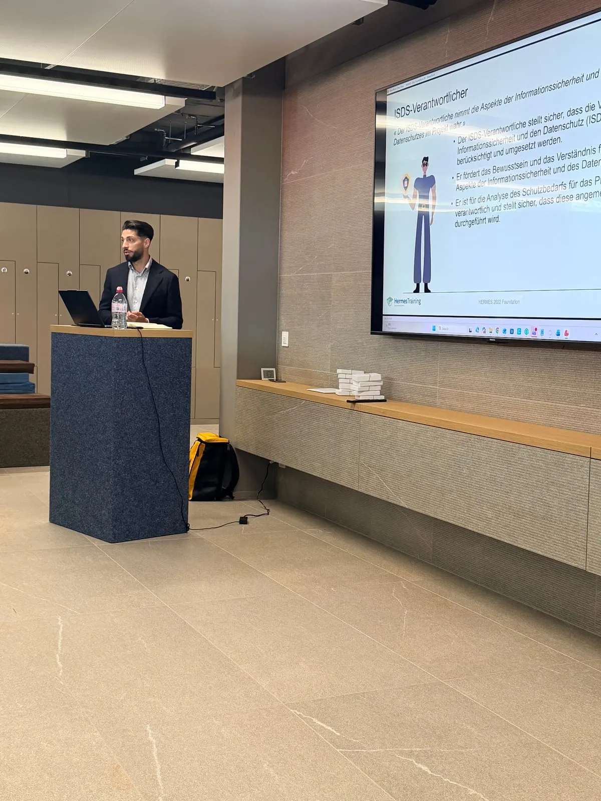 The trainer at the lectern in the xappido arena, next to a large screen showing a HERMES slide on the role of the information security and data protection officer