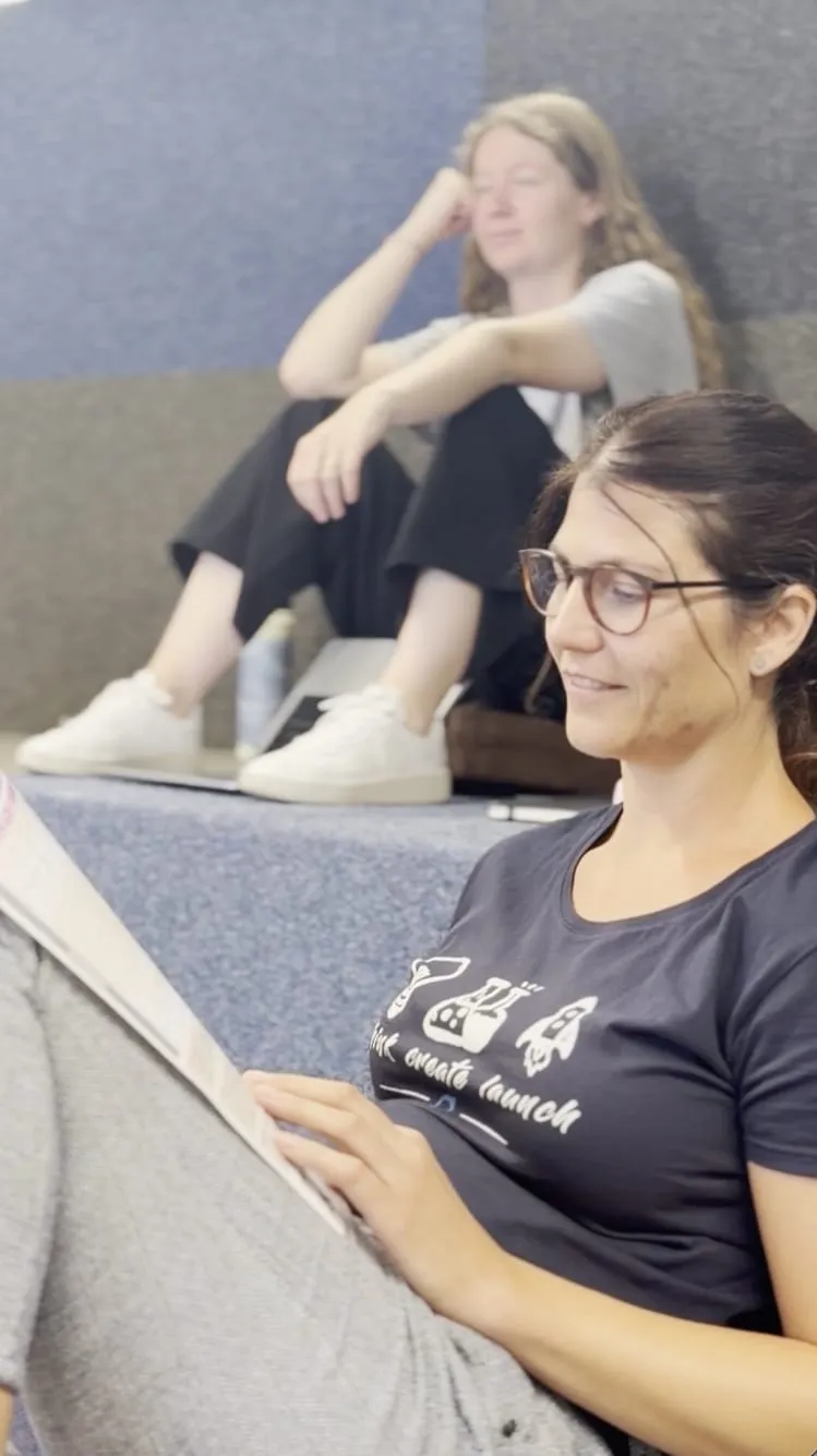 Sandra sitting on the seating steps of the arena, smiling over the course materials, with Fiona behind her