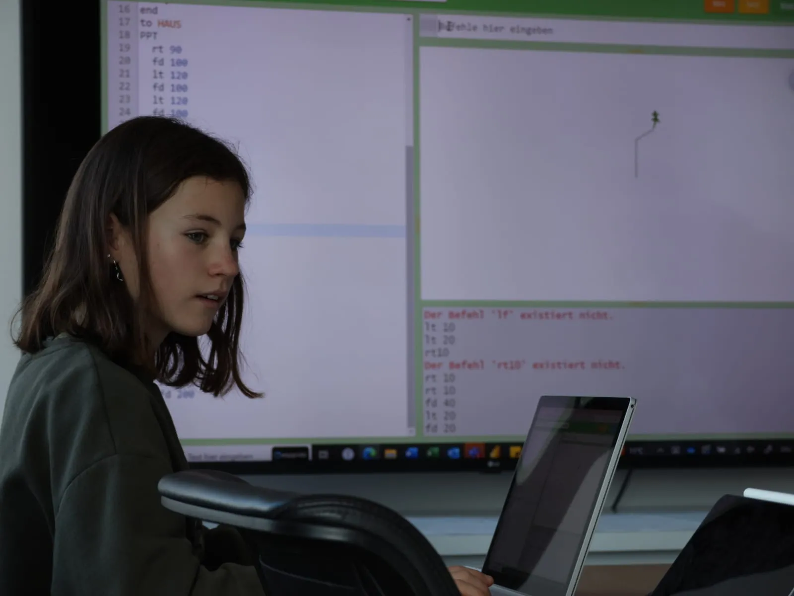 Eine Schülerin am Laptop vor dem grossen Bildschirm mit dem Programmcode