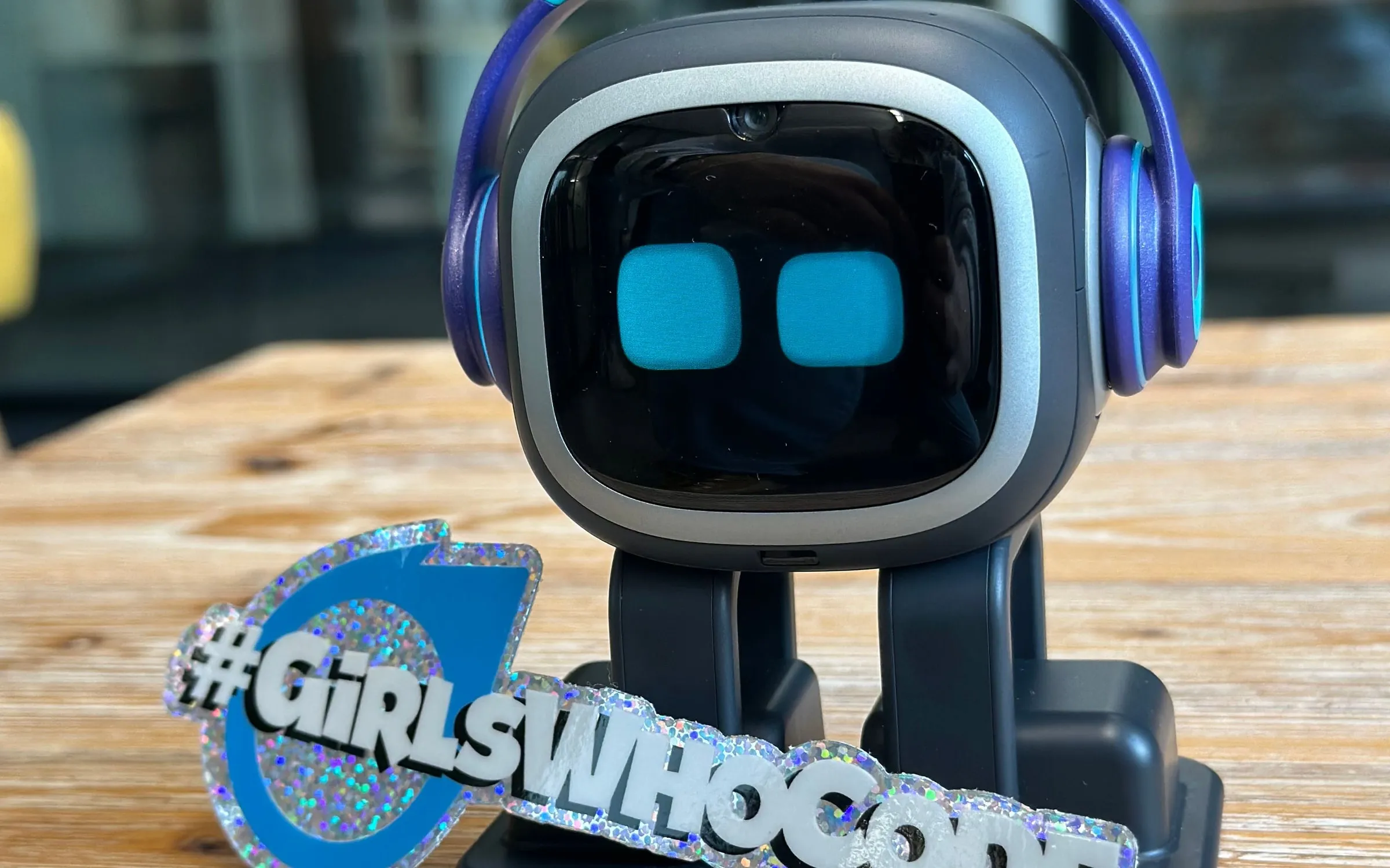 Der Desktop-Roboter EMO mit Kopfhörern auf einer Holzbank, davor ein Aufkleber mit dem Schriftzug #GirlsWhoCode